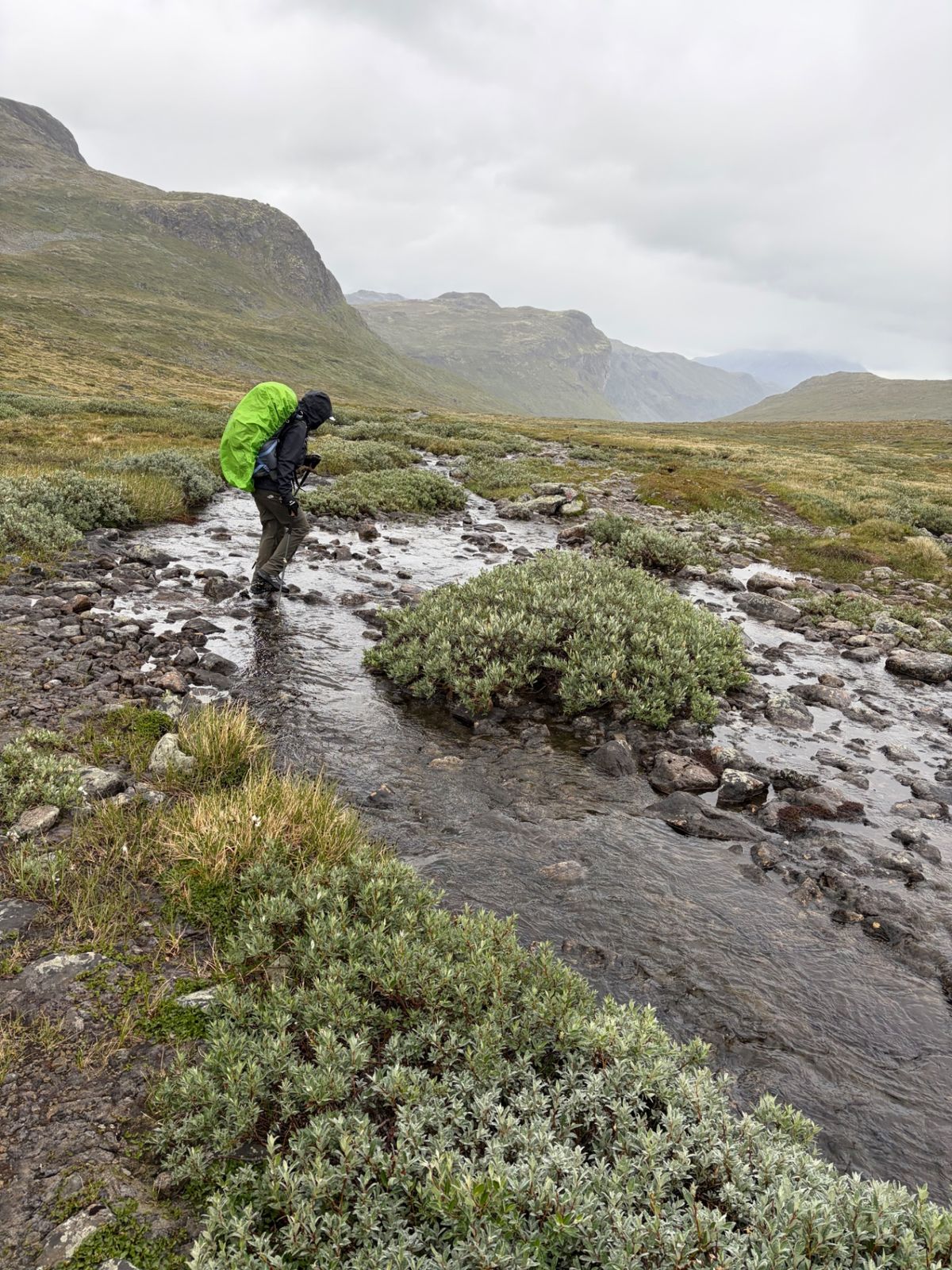 Trekker with a rain cover on the pack fording a stream in the rain