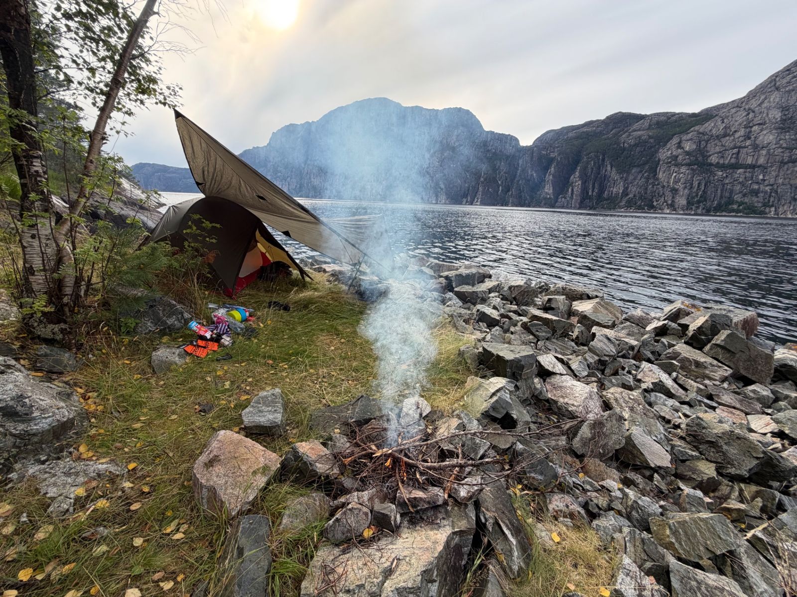 Campfire beside the MSR tent and tarp on the shore of a fjord, steep mountain wall opposite