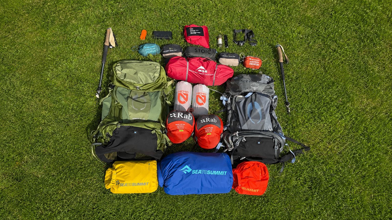 The full set laid out on the grass: two Osprey packs, MSR tent with footprint, Stoic tarp, Rab sleeping bags, NEMO mats, pillows, dry bags, headlamps and trekking poles