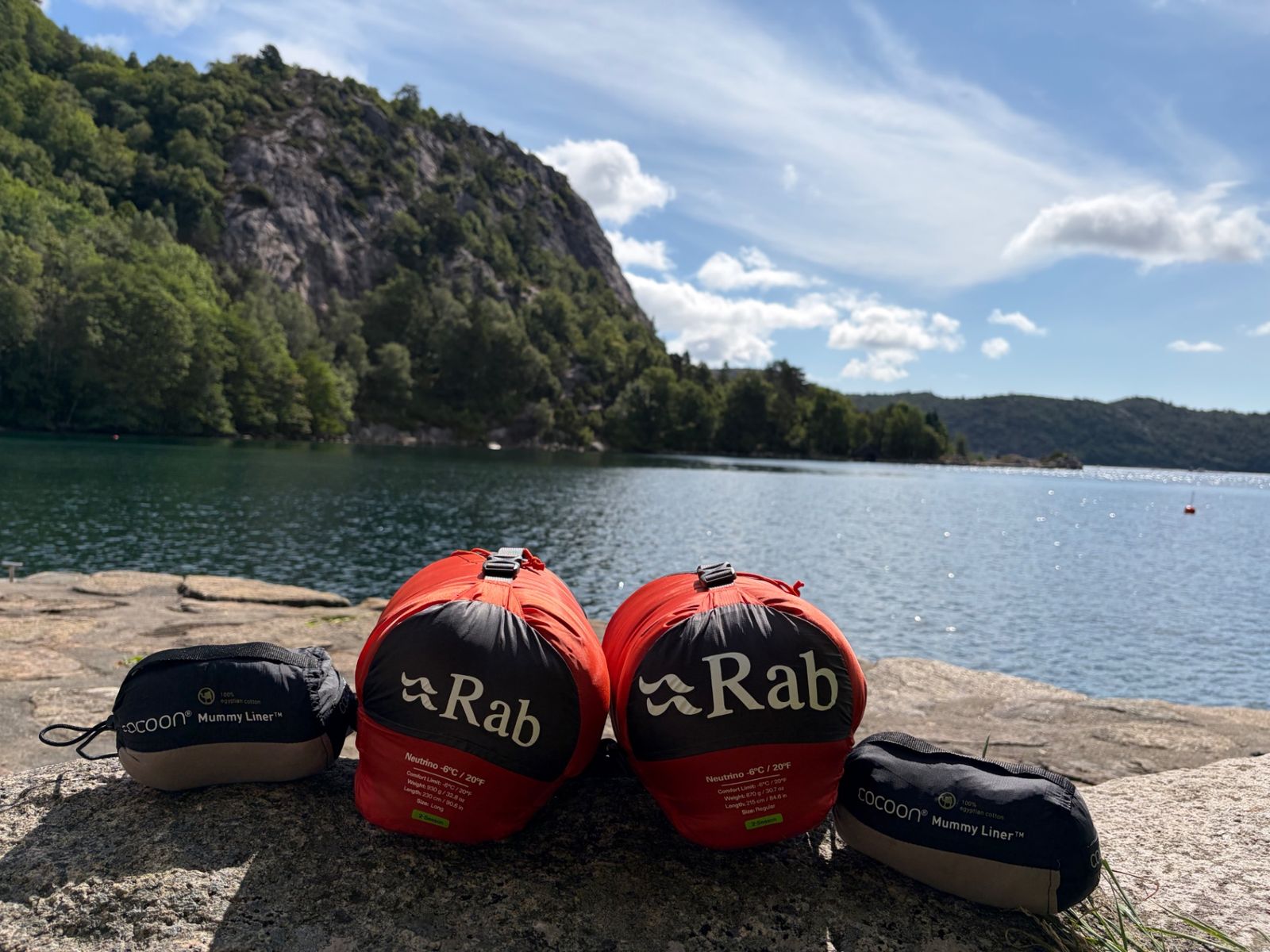 Two Rab Neutrino −6 °C sleeping bags in their red stuff sacks with two Cocoon liners on a rock by the fjord